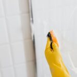 Hand in gele handschoen maakt een glazen douchewand schoon met een spons