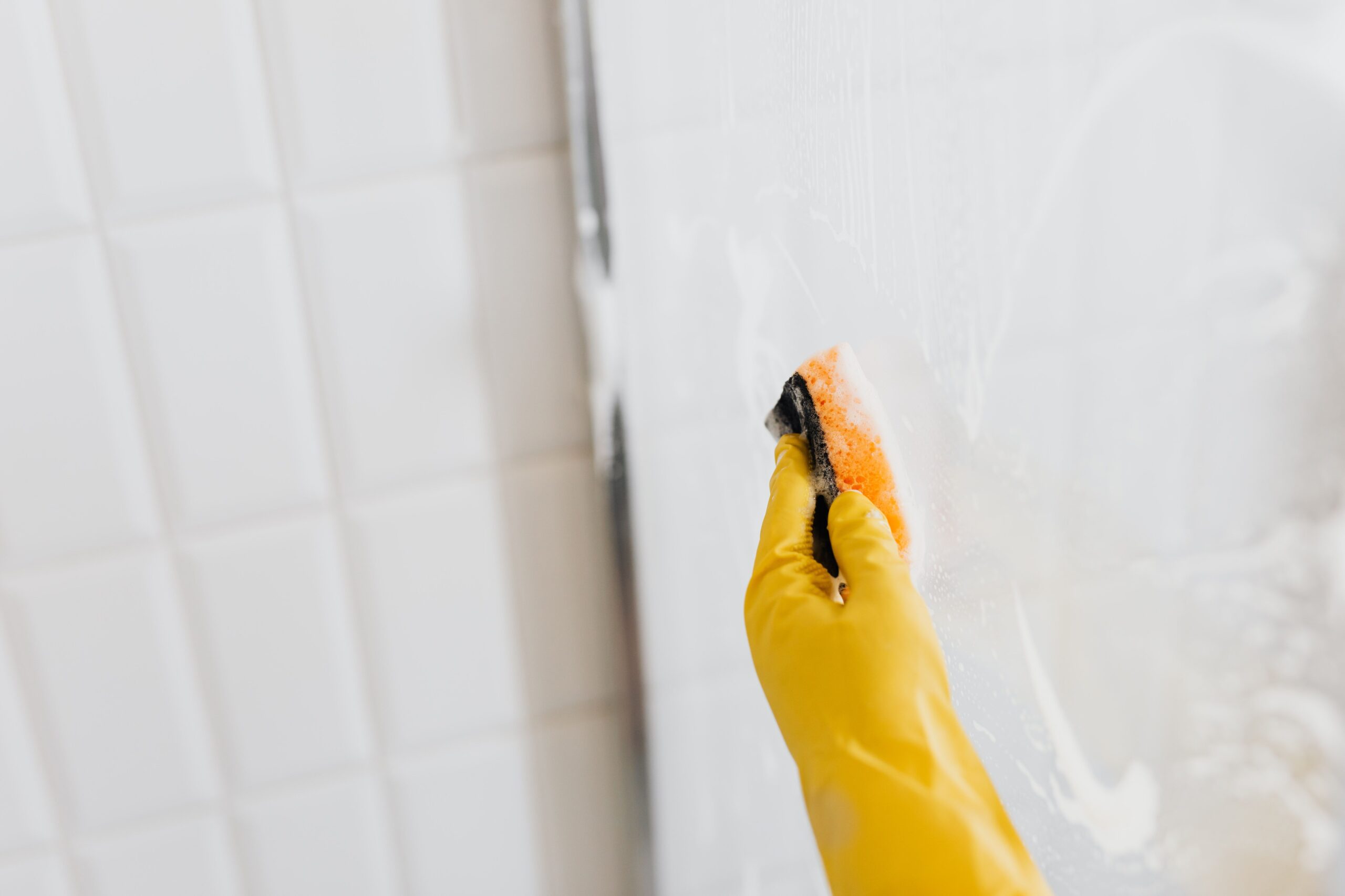 Hand in gele handschoen maakt een glazen douchewand schoon met een spons